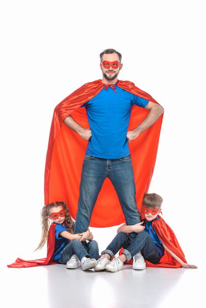 super dad standing with hands on waist and looking at camera while kids hugging his legs isolated on white - Фото, зображення