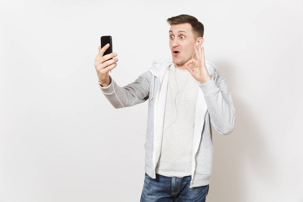Молодой красивый мужчина в футболке и легкой толстовке с наушниками делает селфи на мобильном телефоне во время звонка, показывает жест хорошо изолирован на белом фоне. Концепция общения хорошее настроение
 - Фото, изображение