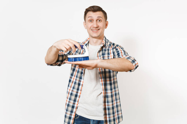Mladý, usměvavý pohledný hospodyně muž v kostkované košili drží modrý kartáč pro čištění izolovaných na bílém pozadí. Muž dělá domácí práce. Kopírovat prostor pro reklamu. Koncept čistoty. - Fotografie, Obrázek