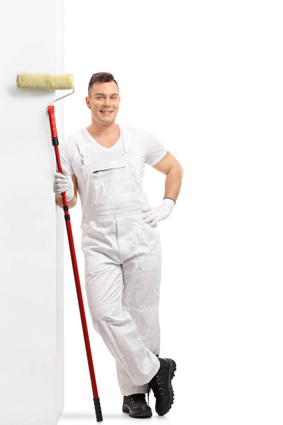Full length portrait of a painter with a paint roller leaning against a wall isolated on white background - Foto, Imagen
