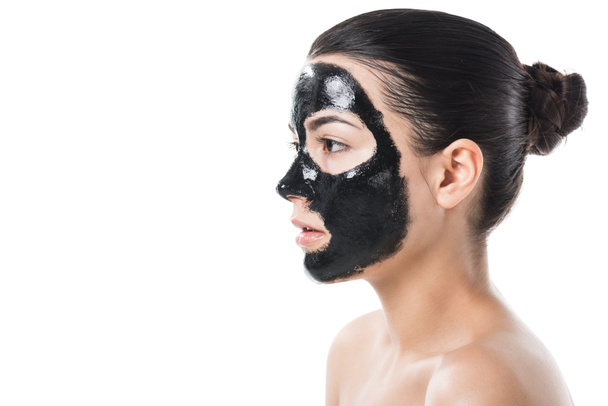 Seitenansicht des brünetten Mädchens in kosmetischer schwarzer Ton Gesichtsmaske isoliert auf weiß - Foto, Bild