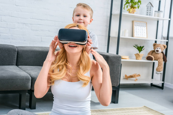 Мати використовує гарнітуру віртуальної реальності з маленьким сином, що стоїть позаду
  - Фото, зображення