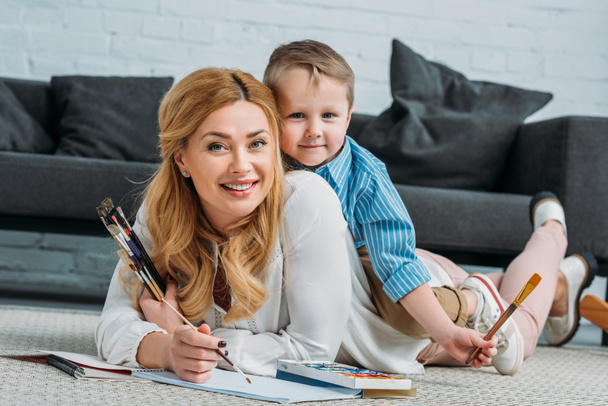 Šťastný malý syn sedí na matku, zatímco ona doma malovat - Fotografie, Obrázek