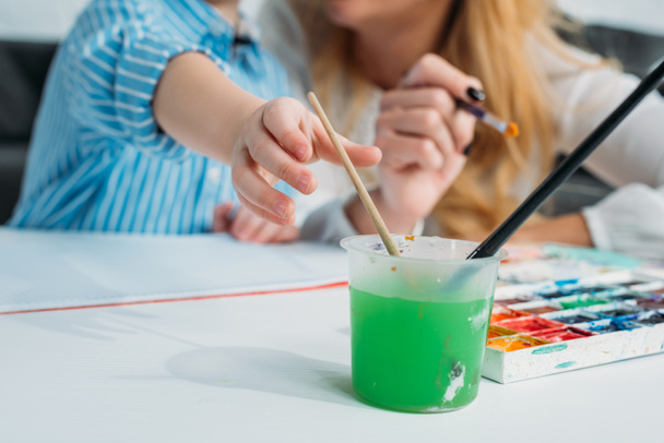 Cropped image of mother and son dipping paintbrush into colored water  - Photo, Image