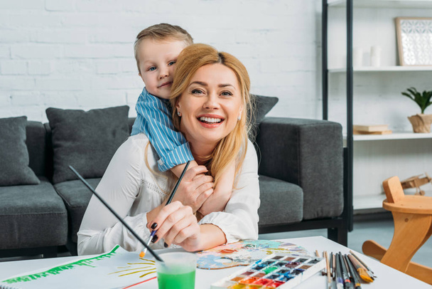 Pequeño hijo abrazando sonriente madre mientras ella pintando en casa
 - Foto, Imagen