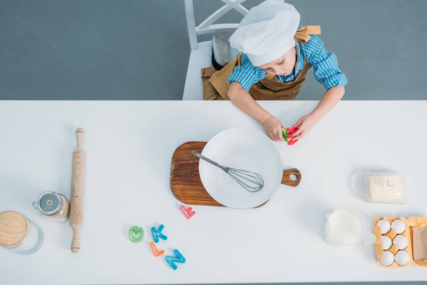 Blick von oben auf den kleinen Jungen mit Kochmütze, der am Tisch mit Geschirr und Zutaten sitzt - Foto, Bild