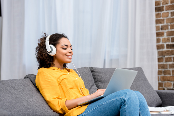 mosolygó afrikai-amerikai nő dolgozik a laptop, és hallgat zenét otthon a kanapén - Fotó, kép