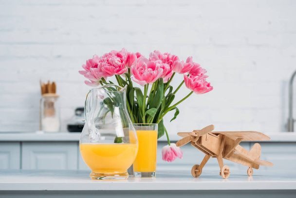 Pink tulips in vase, orange juice and wooden airplane on table in modern kitchen  - Фото, изображение