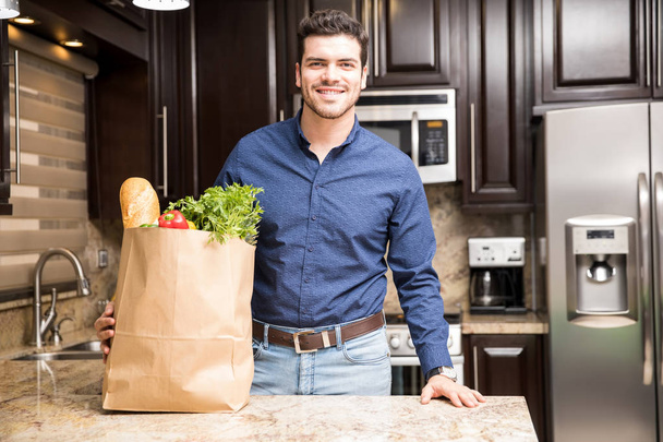 Portrait of handsome young guy standing by kitchen counter with grocery shopping bag full of vegetables - Foto, Bild