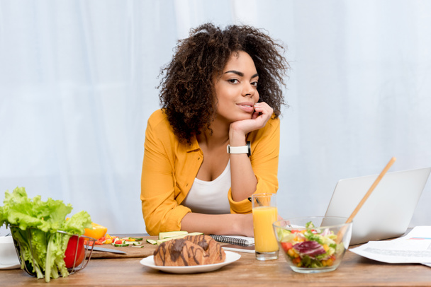 young african american woman leaning on table with various food and laptop, freelance concept - Photo, Image