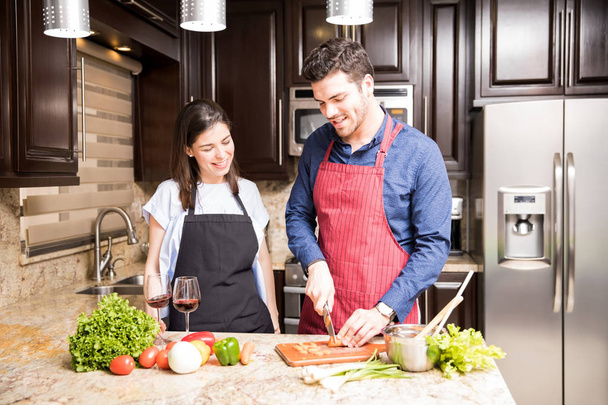 junger lateinischer Mann hackt Gemüse auf Küchentisch, während Frau daneben steht und lächelt - Foto, Bild