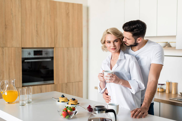 jonge man omhelst vriendin kopje koffie houden en kijken naar camera in keuken - Foto, afbeelding