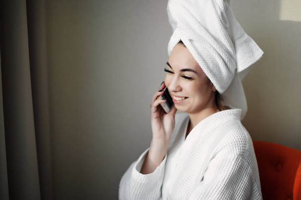 Młoda dziewczyna po prysznicem ze smartfonem owinięta w ręcznik siedzi na krześle w pobliżu okna - Zdjęcie, obraz