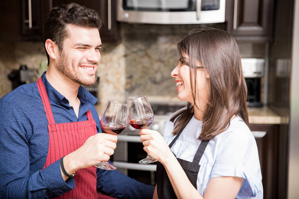 Portrait of happy young couple in apron toasting wine glasses in kitchen - 写真・画像