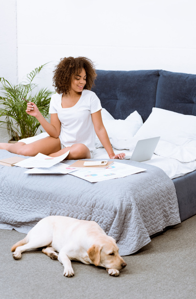 beautiful young woman working at home on bed while her dog lying on floor - Foto, afbeelding
