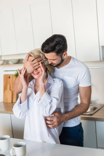 handsome young man closing eyes to smiling girlfriend in kitchen - Foto, Bild