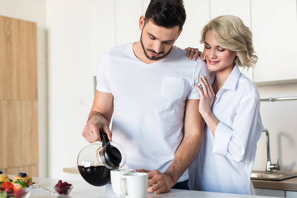 smiling young woman hugging handsome boyfriend while he pouring coffee in kitchen - Foto, afbeelding