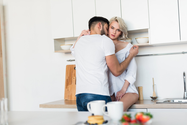 young man hugging and kissing sexy girlfriend, girl looking at camera in kitchen - Photo, Image