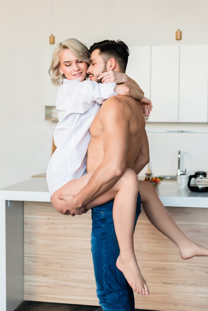 schöner hemdloser junger Mann trägt und umarmt sexy Freundin in der Küche  - Foto, Bild