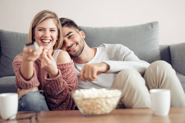 young couple sitting near sofa at home with popcorn and watching TV - Photo, Image