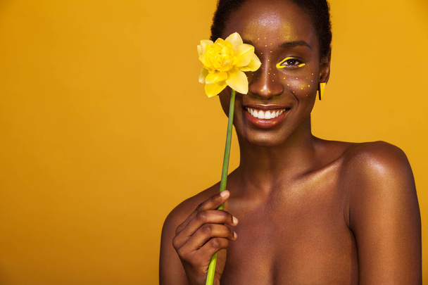 Cheerful young african woman with yellow makeup on her eyes. Female model laughing against yellow background with yellow flower. - Fotó, kép