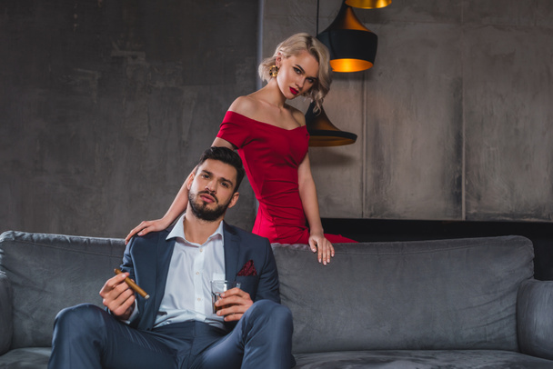 man in suit holding cigar and glass of whisky while spending time with sexy girl in red dress - Fotó, kép