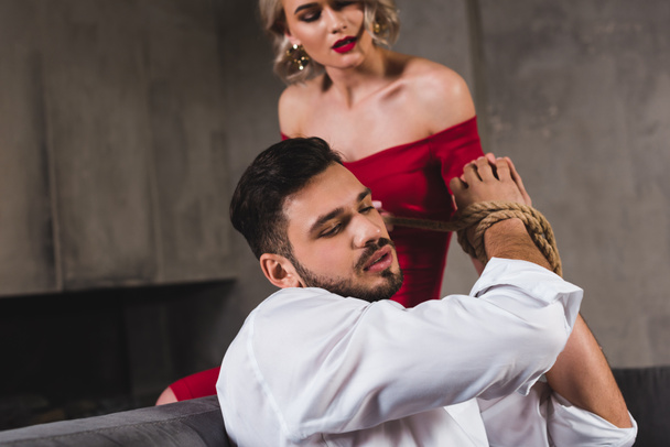 sensual young woman in red dress tying hands of handsome boyfriend with rope  - Foto, afbeelding