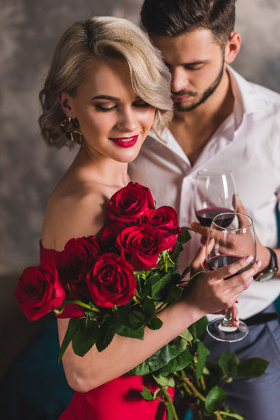 smiling young woman in red dress holding beautiful roses and drinking wine with handsome boyfriend  - Zdjęcie, obraz