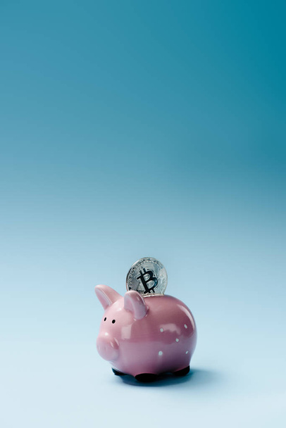 Close up view of pink piggy bank and silver bitcoin on blue backdrop - Zdjęcie, obraz