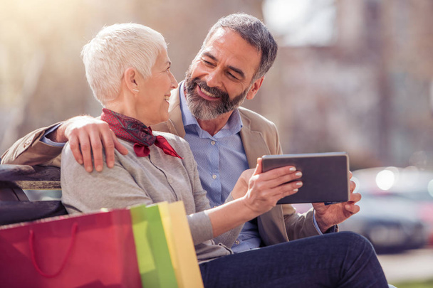 Älteres Paar sitzt mit Einkaufstaschen und Tablet-Computer auf Bank - Foto, Bild
