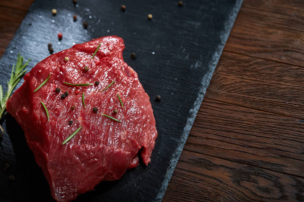 Raw meat beef steaks on black slate board with spices and rosemary over wooden background, copy space - 写真・画像