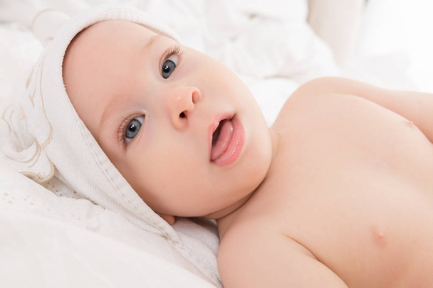 Happy baby boy wearing hooded towel lying on a bed after bath or shower. Young child with blue eyes looking at camera. - Foto, afbeelding