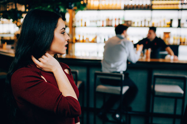 Beautiful picture of attractive brunette sitting not very far from the barmans stand. She is looking to the side where are barman and his customer. She looks excited. - Photo, Image