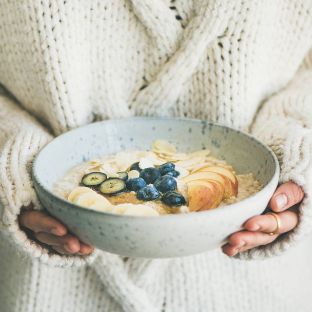 Healthy winter breakfast in bed. Woman in woolen sweater holding bowl of vegan almond milk oatmeal porridge with berries, fruit and almonds - Фото, зображення