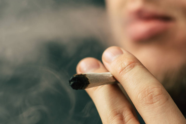男は、彼の手で大麻雑草、ジョイントとライターを吸います。黒の背景にたばこを吸います。医療マリファナの使用と大麻の合法化の概念. - 写真・画像