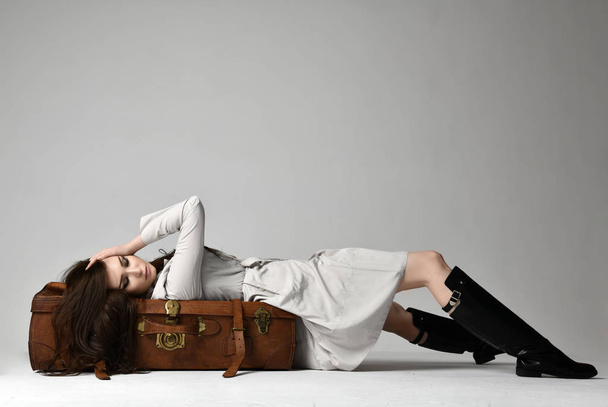 Beautiful brunette hipster woman in gray autumn jacket lying on leather brown retro travel bag on grey - Valokuva, kuva