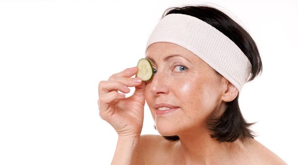 Cheerful mature woman holding piece of cucumber in front of her eye - Foto, afbeelding