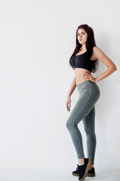 Full-length side portrait of the young athletic woman in black short top and gray leggings posing at white background. - Photo, image