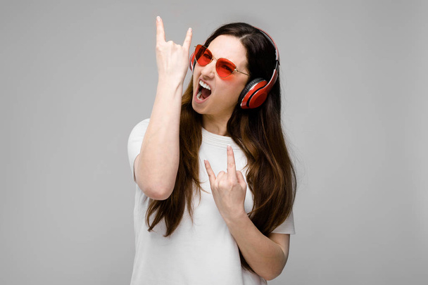 Beautiful curvy brunette woman with long luxurious hair in simple white t-shirt, in red sunglasses and red headphones showing different expressions on grey wall in studio. - Foto, Imagem