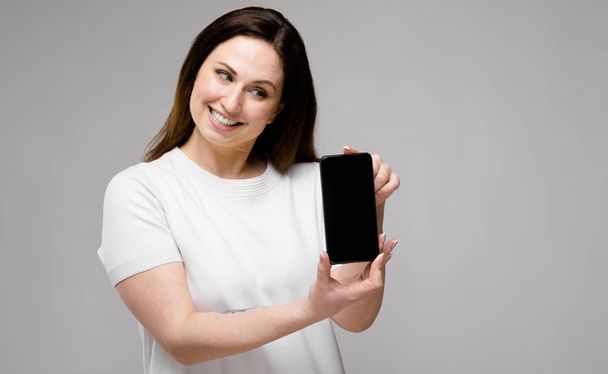 Beautiful curvy brunette woman with long luxurious hair in simple white t-shirt showing different expressions on grey wall and showing blank screen of smartphone - Foto, Imagen