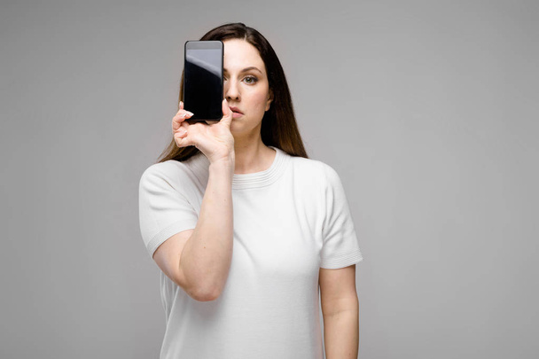 Beautiful curvy brunette woman with long luxurious hair in simple white t-shirt showing different expressions on grey wall in studio while posing with her smartphone. - Foto, immagini