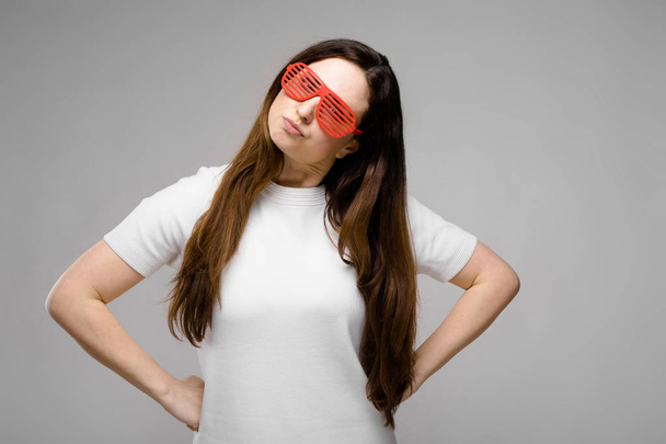 schöne kurvige brünette Frau mit langen luxuriösen Haaren in einfachem weißen T-Shirt und roter Party-Sonnenbrille aus Plastik, die verschiedene Ausdrucksformen an grauer Wand im Studio zeigt. - Foto, Bild