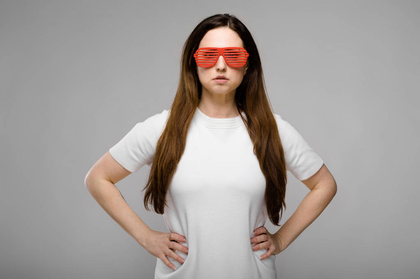 Belle femme brune courbée avec de longs cheveux luxueux en simple t-shirt blanc et lunettes de soleil de fête en plastique rouge montrant différentes expressions sur le mur gris en studio
. - Photo, image