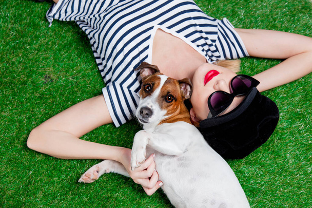 Zrzka dívka v sluneční brýle a klobouk s psa vleže na zelené trávě. - Fotografie, Obrázek