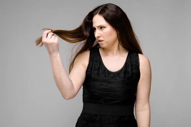 Krásná curvy bruneta žena s dlouhou luxusní vlasy v jednoduché černé tričko ukazující různé výrazy na šedou stěnu ve studiu. Při pohledu na konečky vlasů se zklamání. - Fotografie, Obrázek
