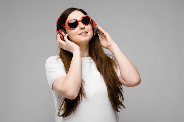 シンプルな白の t シャツで髪の長い豪華な美しい曲線のブルネットの女性、灰色のさまざまな表現を示す赤いヘッドフォンでハート形の眼鏡壁のスタジオで. - 写真・画像