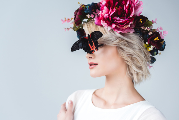 stylish girl posing in floral wreath with beautiful alive butterfly on face, isolated on grey - Foto, afbeelding