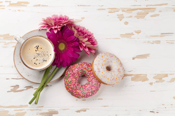 Xícara de café, donuts e flores de gerbera na mesa de madeira branca. Vista superior com espaço para o seu texto
 - Foto, Imagem