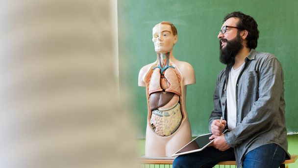 Young male hispanic teacher in biology class, holding digital tablet and teaching human body anatomy, using artificial body model to explain internal organs. - Photo, image