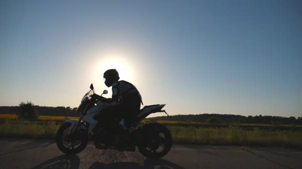 Man in helmet riding on a motorbike at highway with sun flares at background. Motorcyclist driving his motorcycle on country road during sunset. Slow motion Side view Close up - Footage, Video